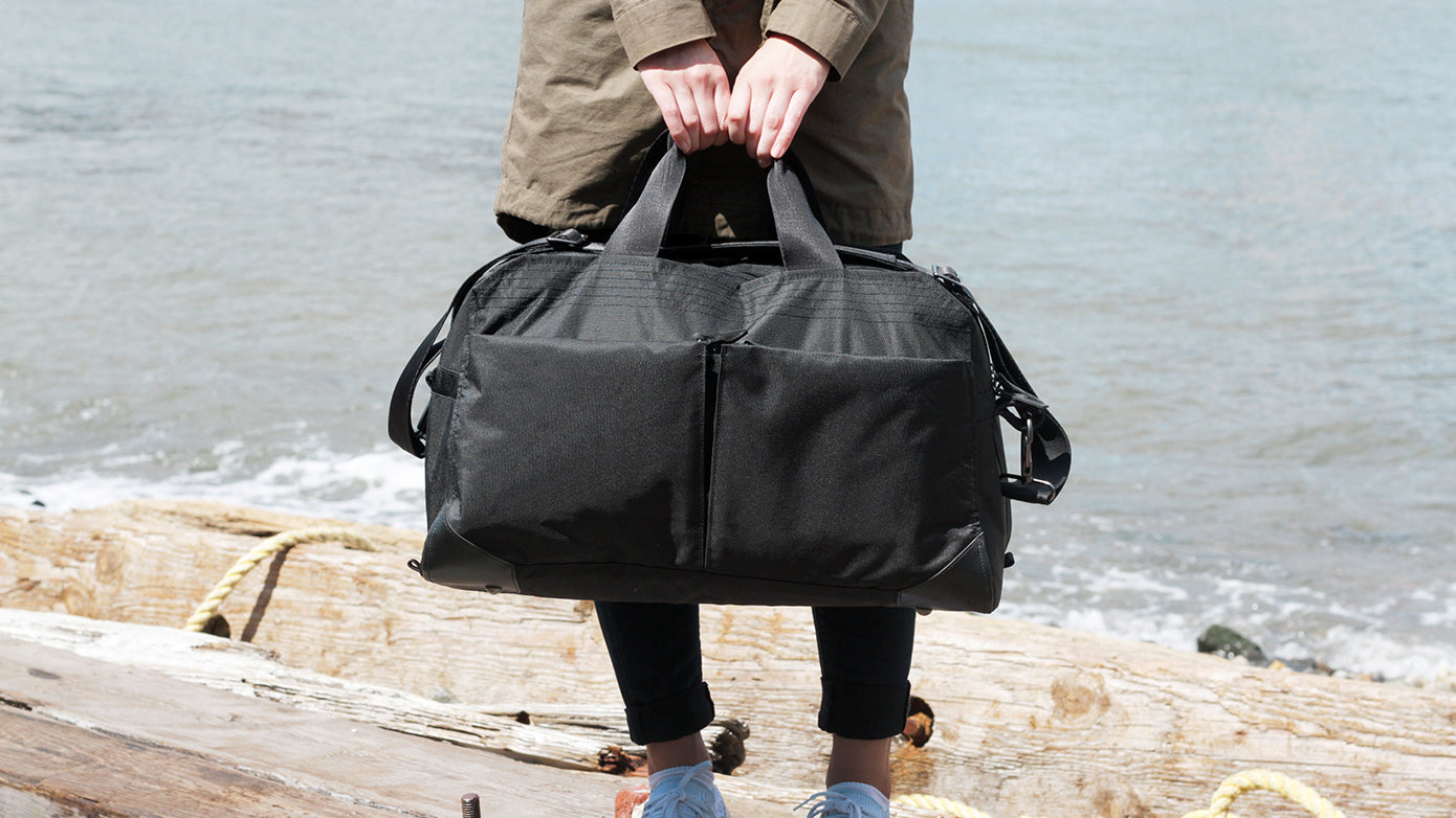 Person holding a black V1 Grandcart One Duffel bag by the water's edge, standing on wooden logs.