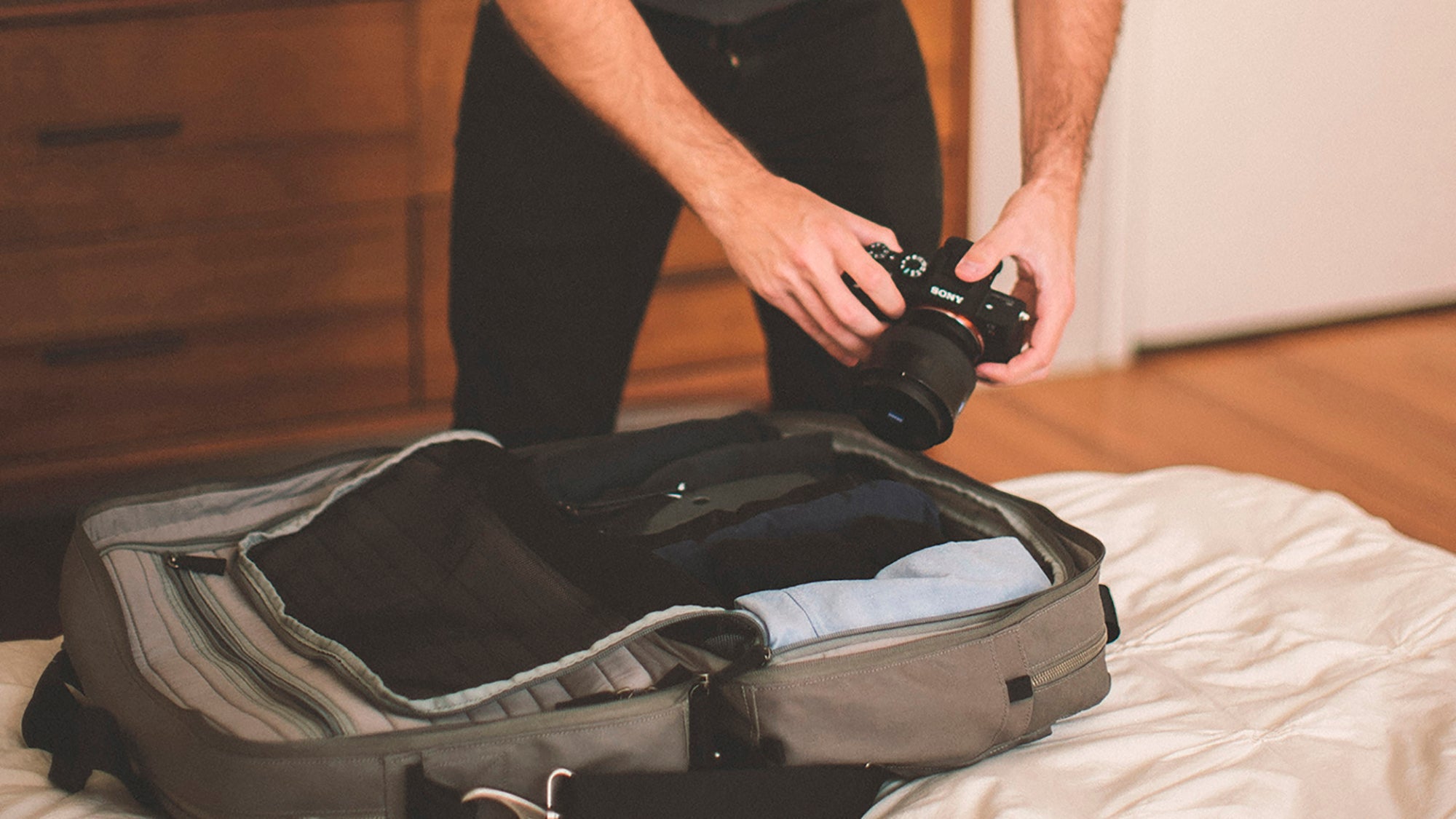 Person holding a Sony camera over an open Grandcart One with folded clothes on a bed in a room