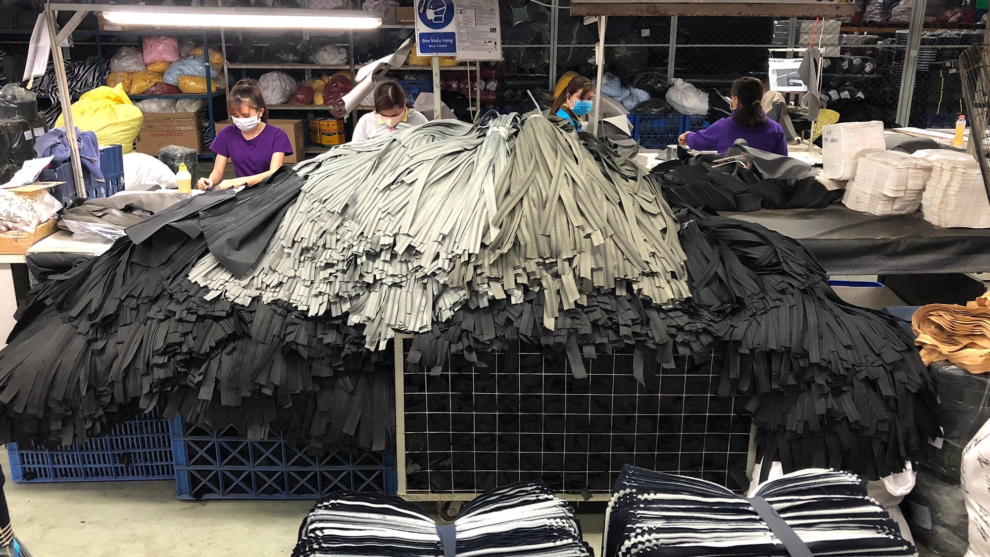 Four masked workers sorting large piles of black and gray fabric strips in a textile factory workspace.