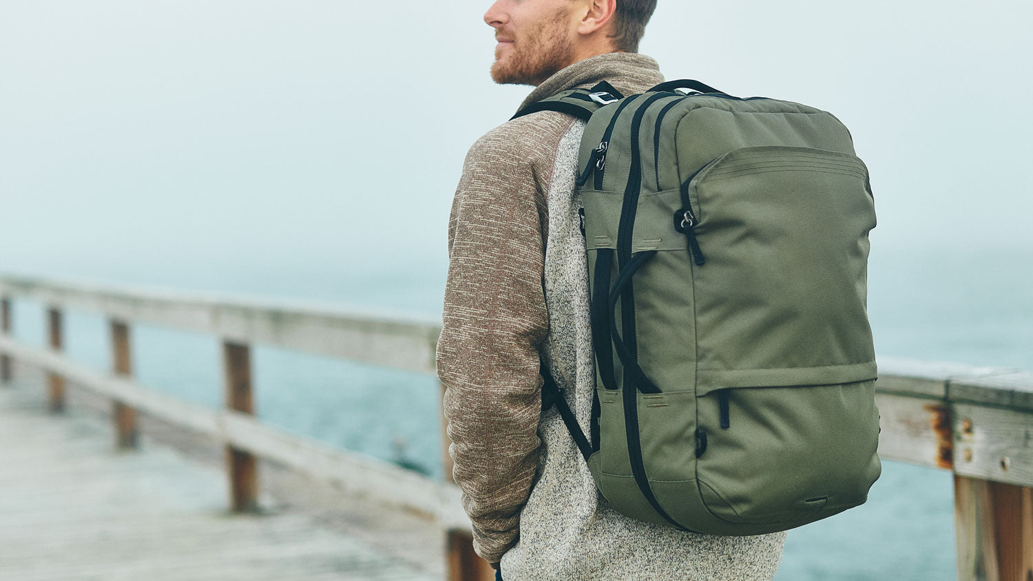 Man wearing a large V1 green Travel Backpack standing on a wooden pier by the water, facing sideways.