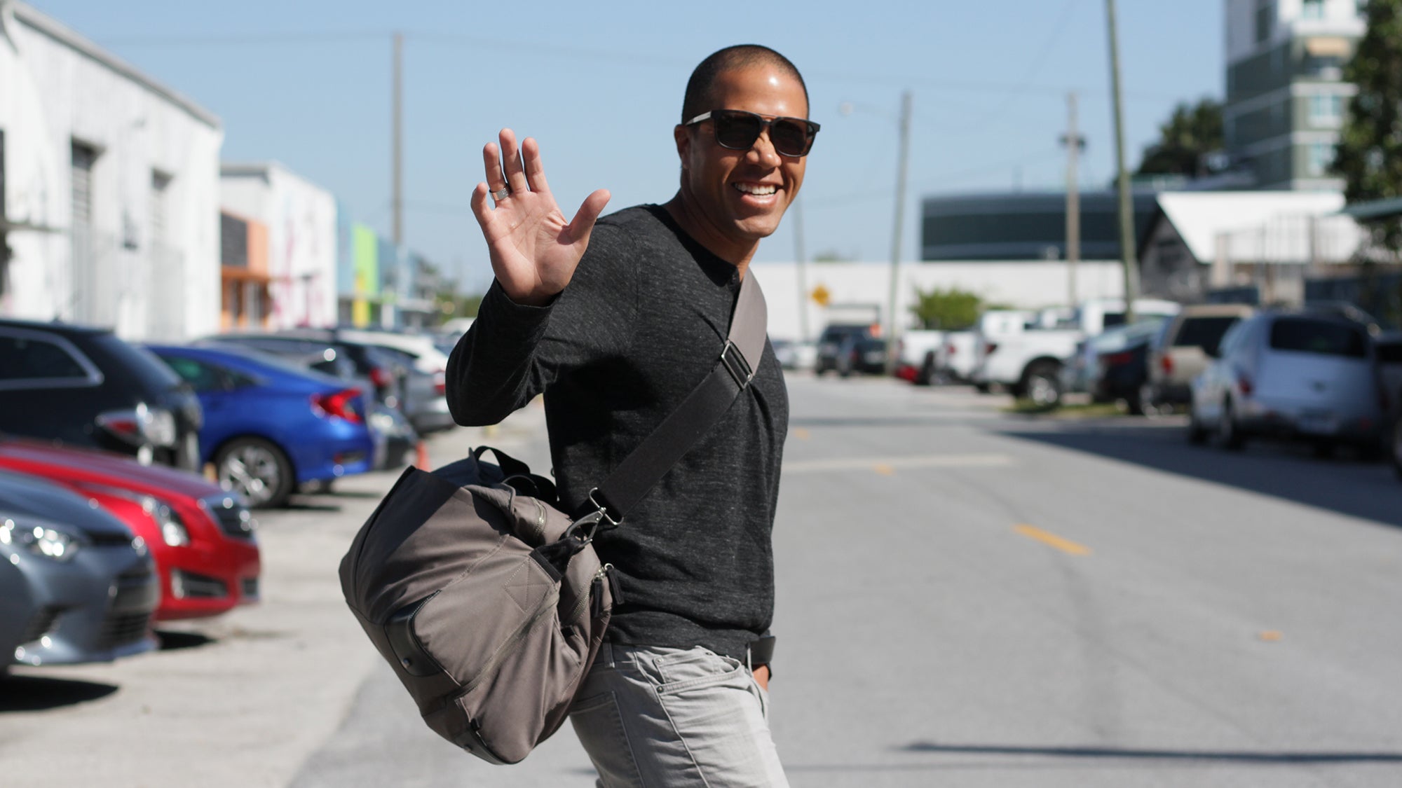 Smiling man wearing sunglasses waving with his right hand, carrying a gray V1 Grandcart One bag, standing on a street with parked cars and buildings in the background.