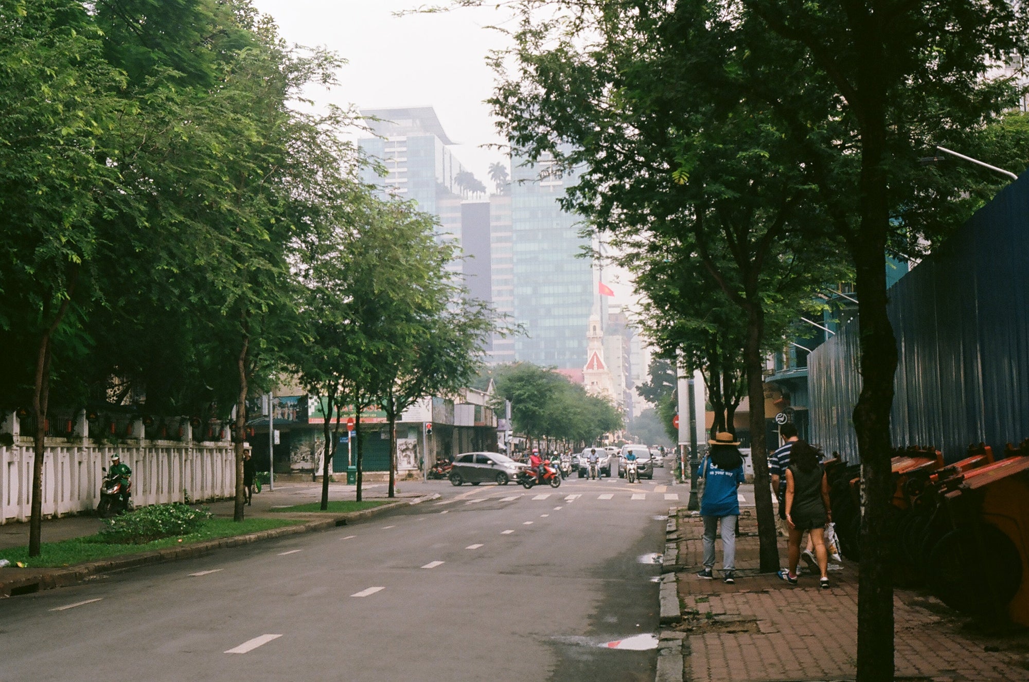 City street lined with trees, pedestrians walking on the sidewalk, and vehicles in the distance under an overcast sky.