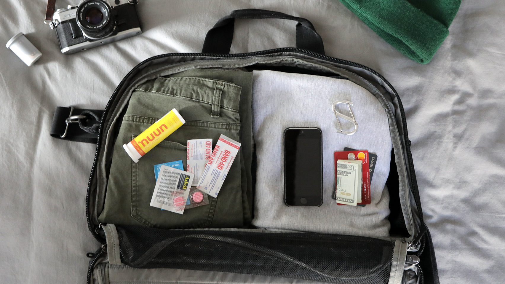 An open V1 Grandcart One Travel Bag with folded green pants, gray shirt, smartphone, cash, credit cards, band-aids, medication, and a carabiner clip on a bed.
