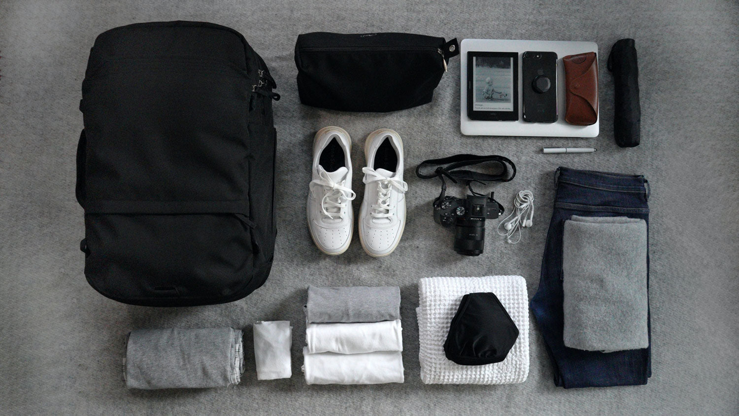 A Black V1 Travel Backpack, black pouch, white sneakers, folded grey and white clothes, camera with strap, earphones, folded jeans with grey sweater, tablet, smartphone, sunglasses case, pen, and folded umbrella arranged on grey carpet.