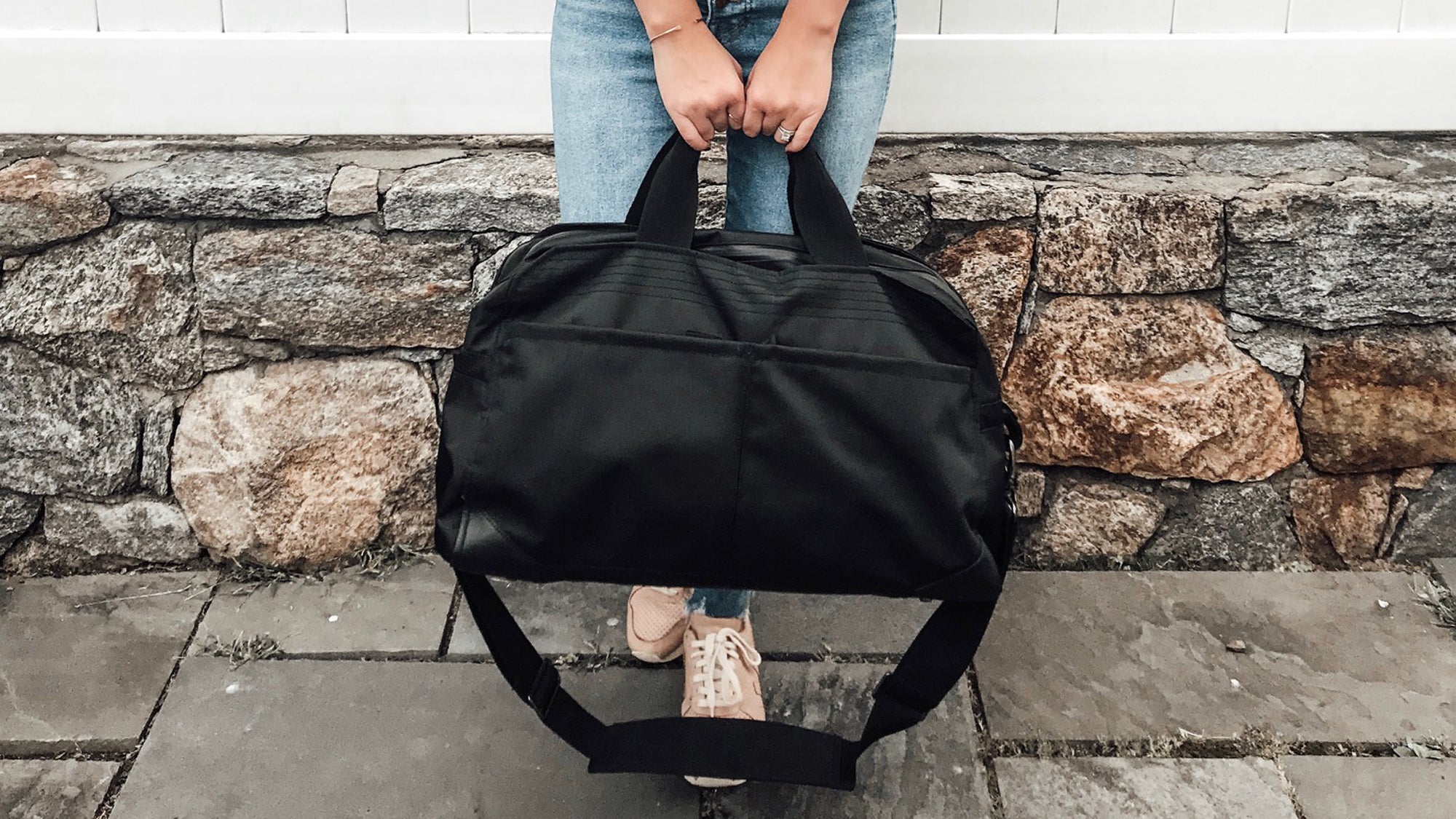Person holding a black Grandcart One travel bag by its handles, standing on stone pavement in front of a stone wall.