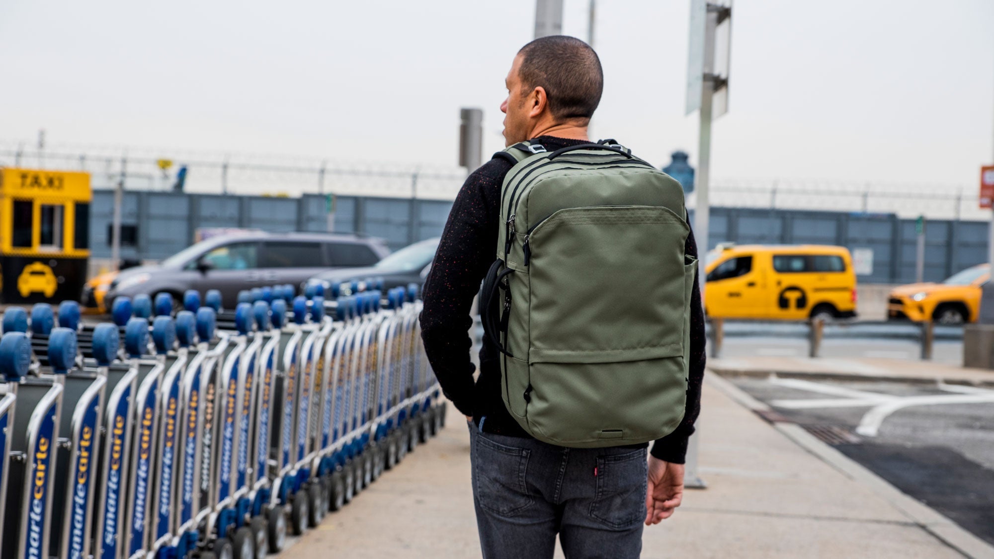 Man walking on sidewalk wearing the V1 Travel Backpack near luggage carts and yellow taxis