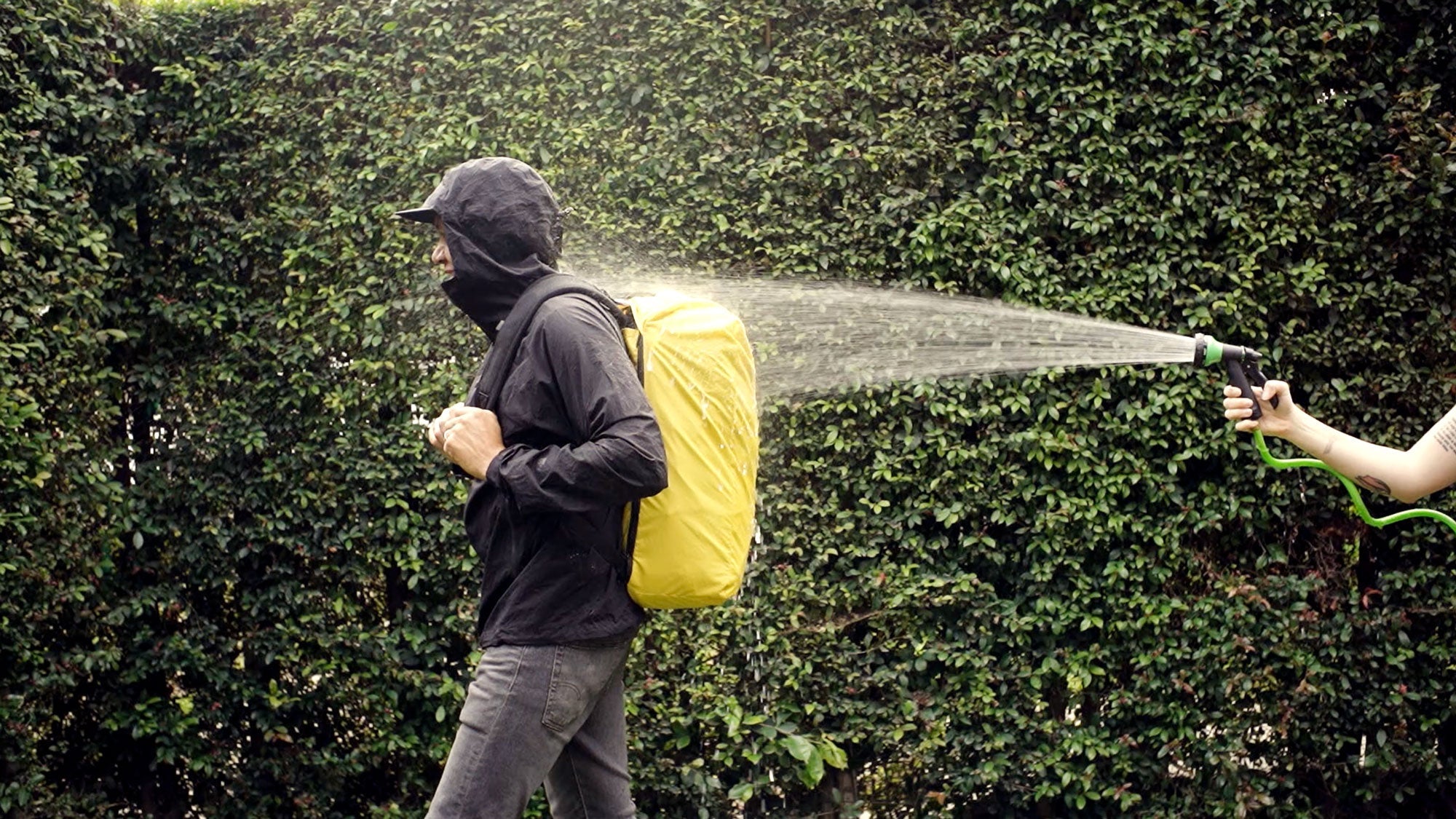 Person wearing a yellow backpack being sprayed with water from a garden hose, demonstrating water resistance