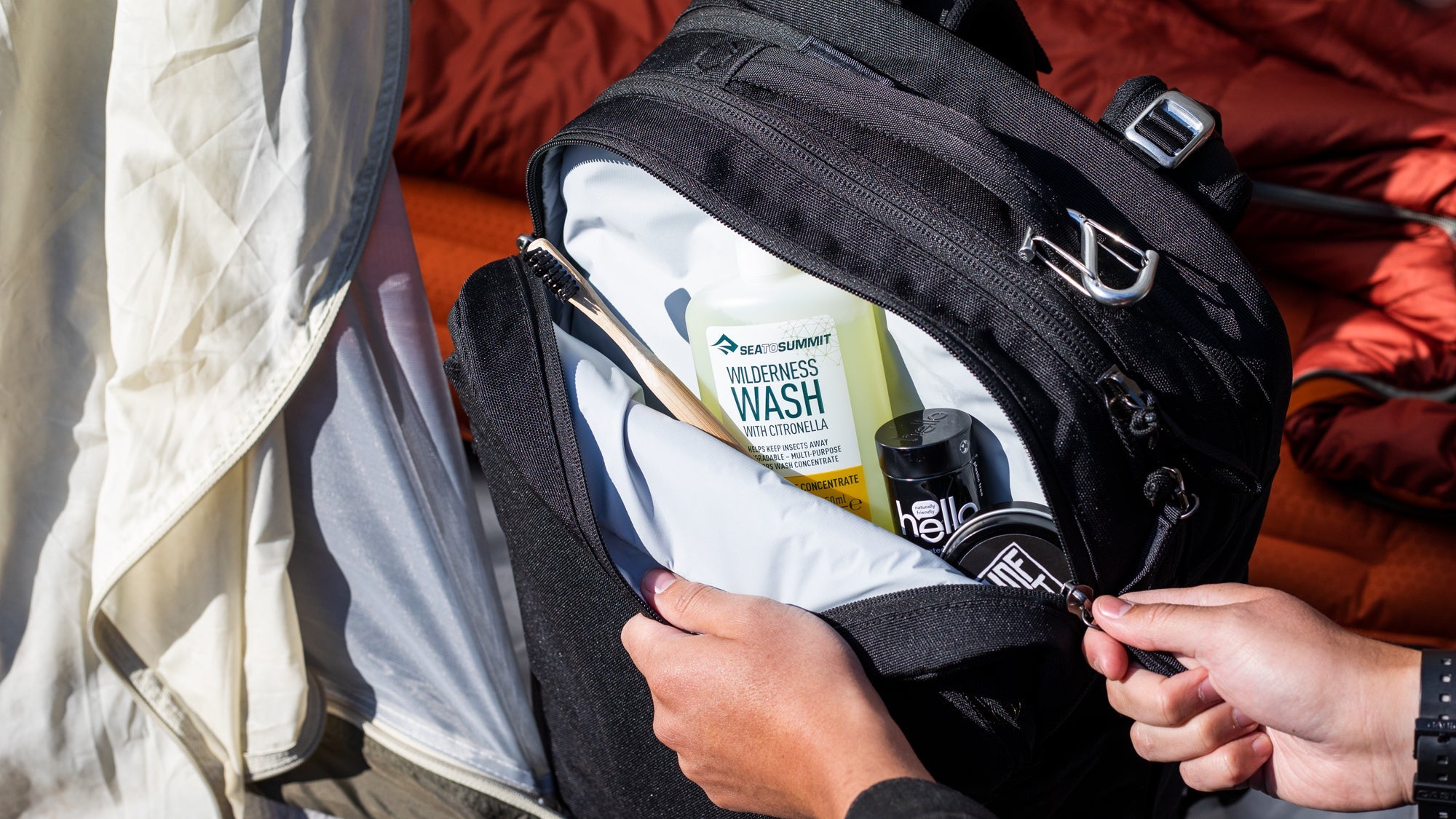 Hands opening a black travel bag containing a bamboo toothbrush, a bottle labeled 'SEA TO SUMMIT WILDERNESS WASH WITH CITRONELLA', a black container labeled 'hello', and a round tin labeled 'WTF'.