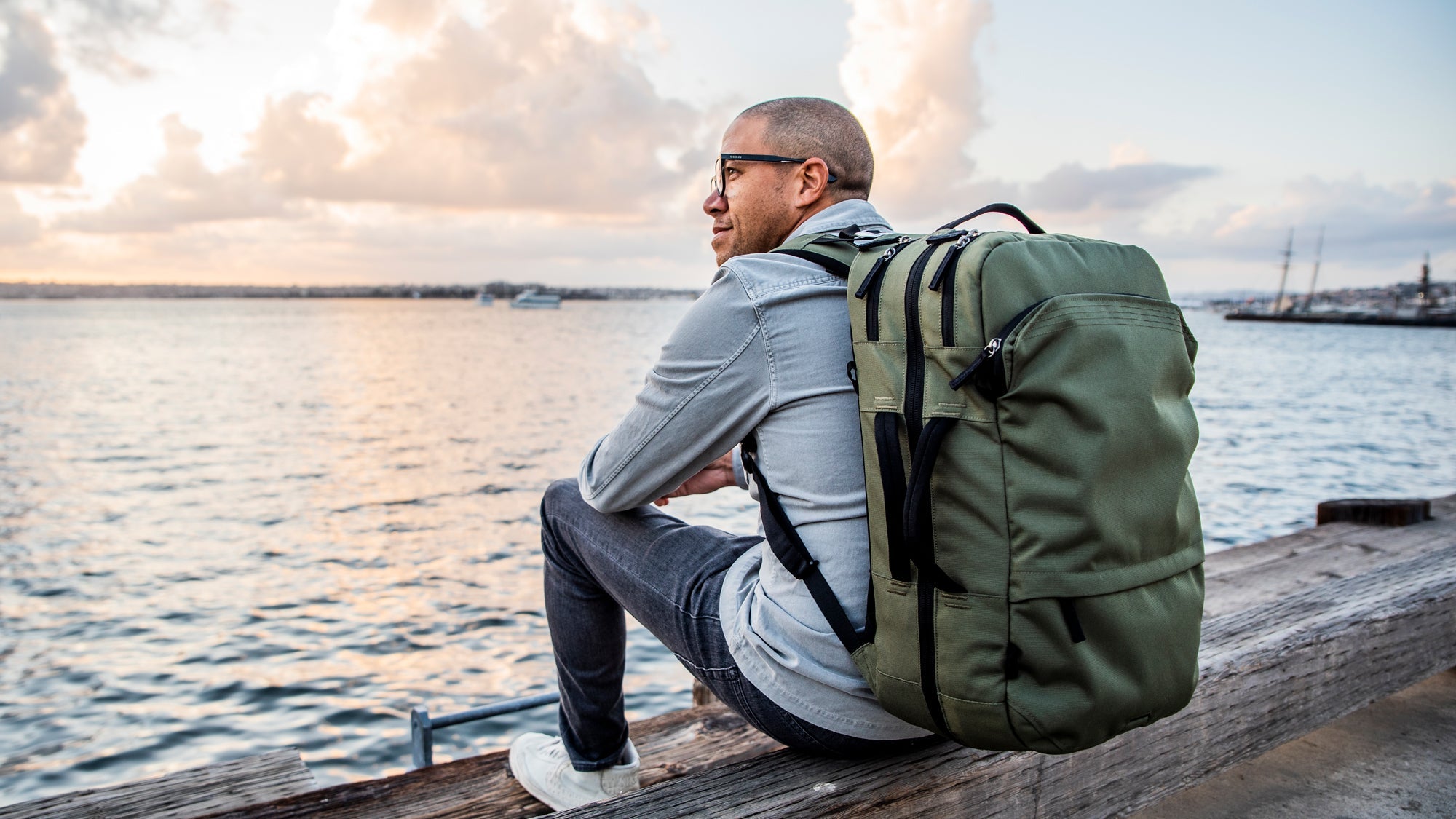 Man with glasses sitting on a wooden dock by the water at sunset, wearing a large green backpack, looking contemplative.