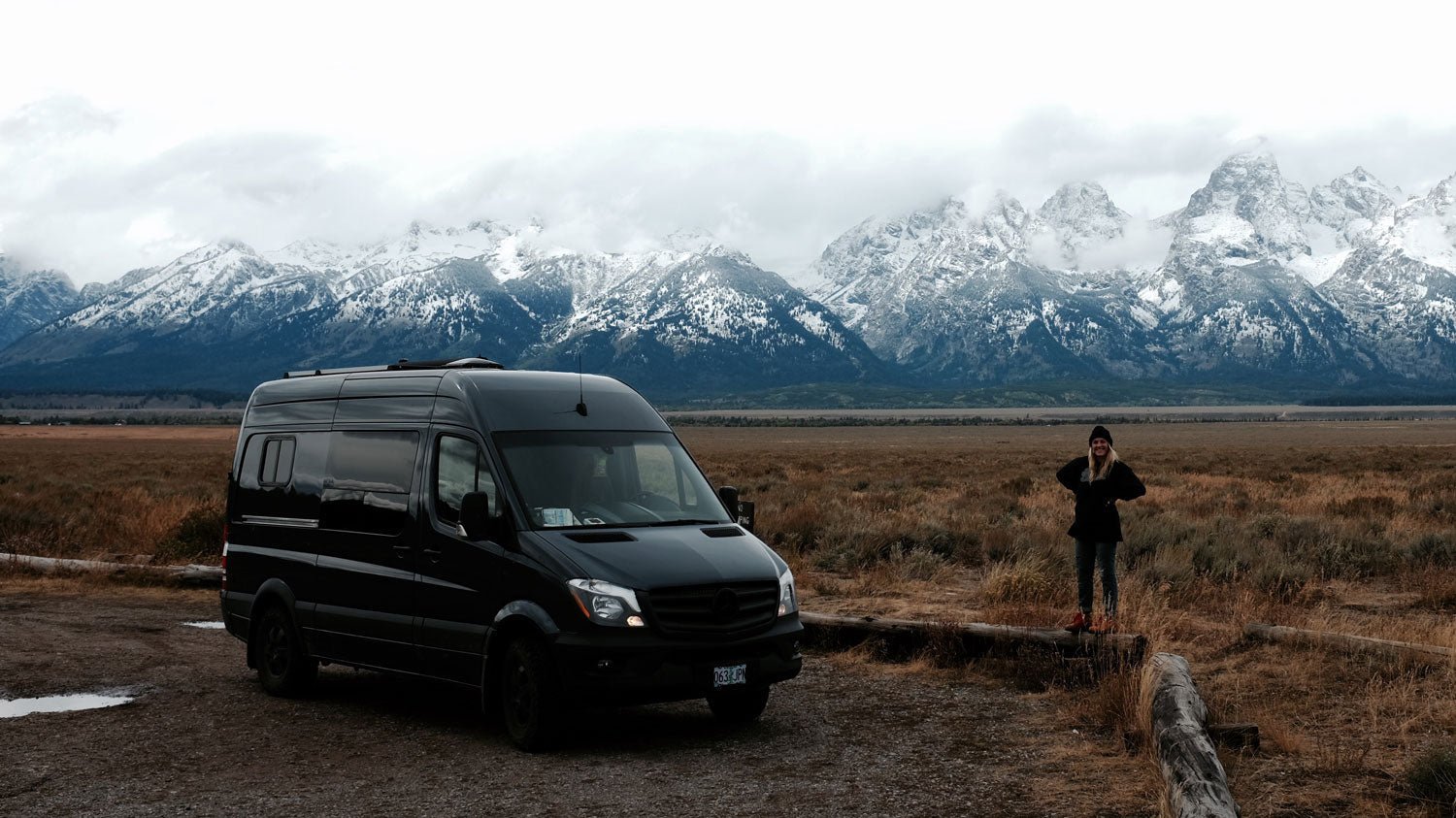 Black camper van parked on a dirt road with a person standing nearby in a field, snowy mountains in the background under a cloudy sky.