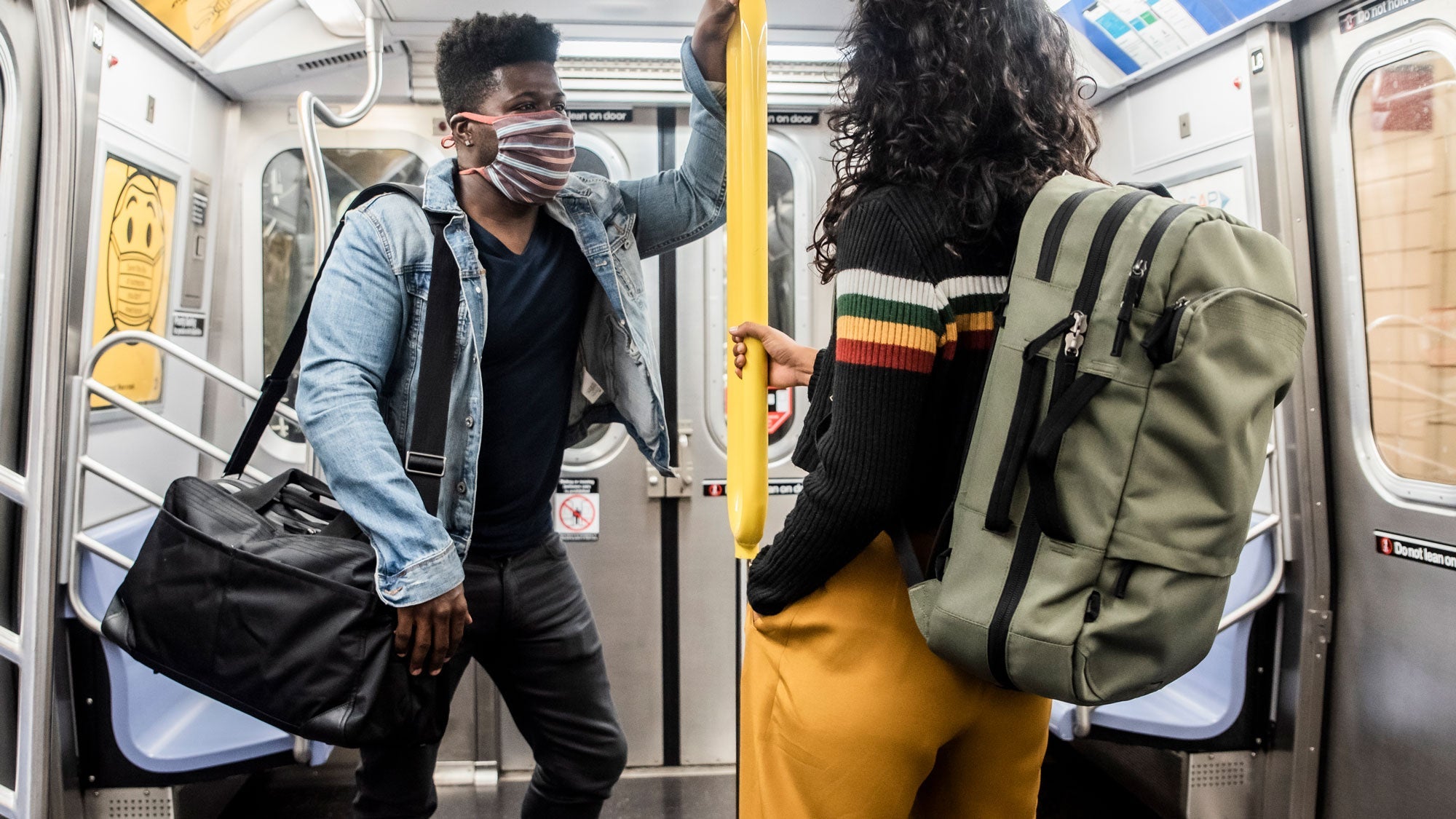 Two people wearing masks on a subway, one carrying a black duffel bag and the other wearing a green backpack, standing and holding a yellow pole.