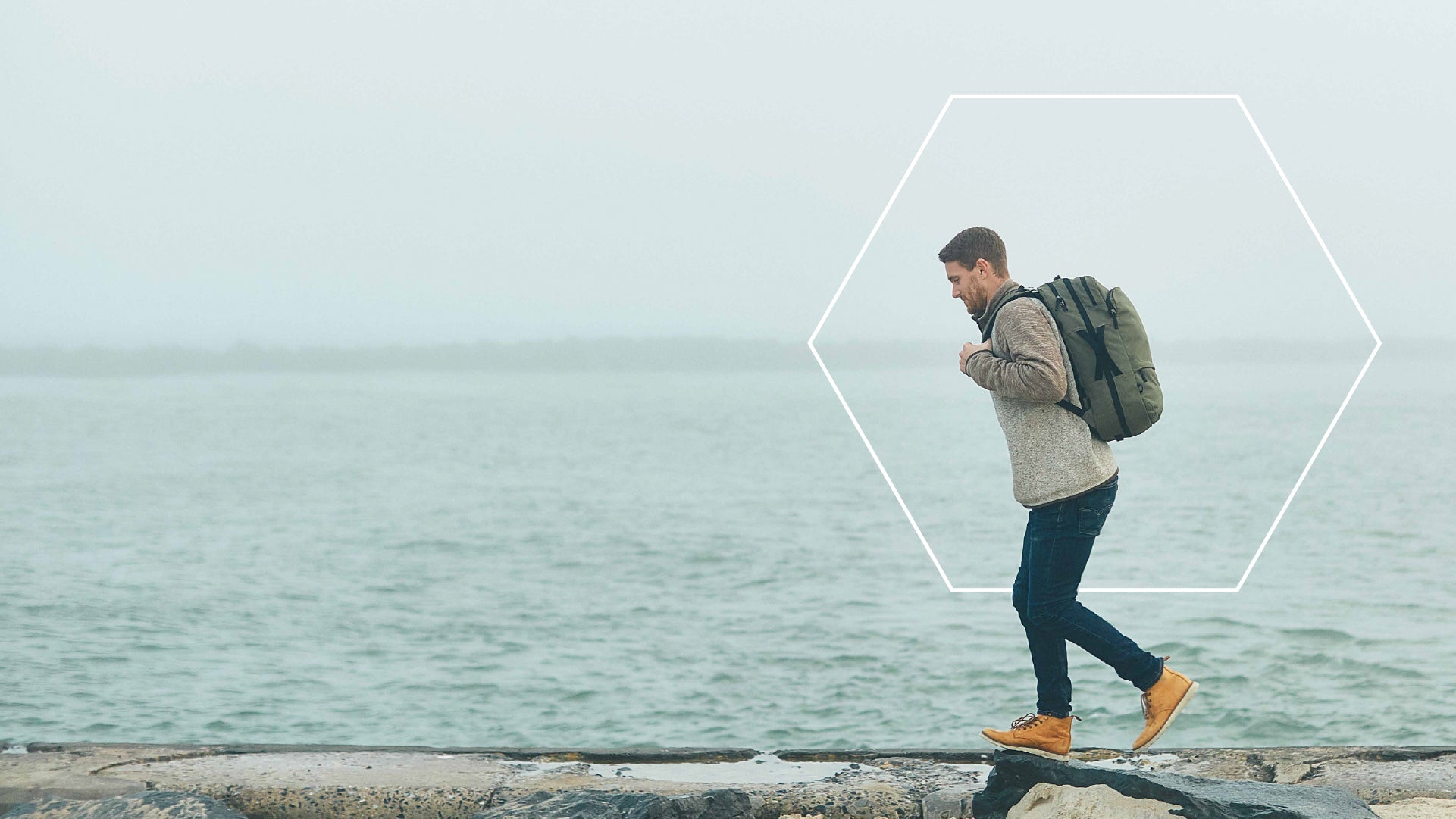 Man walking on rocks by the water carrying a green V1 Travel Backpack with a hexagonal outline around him.