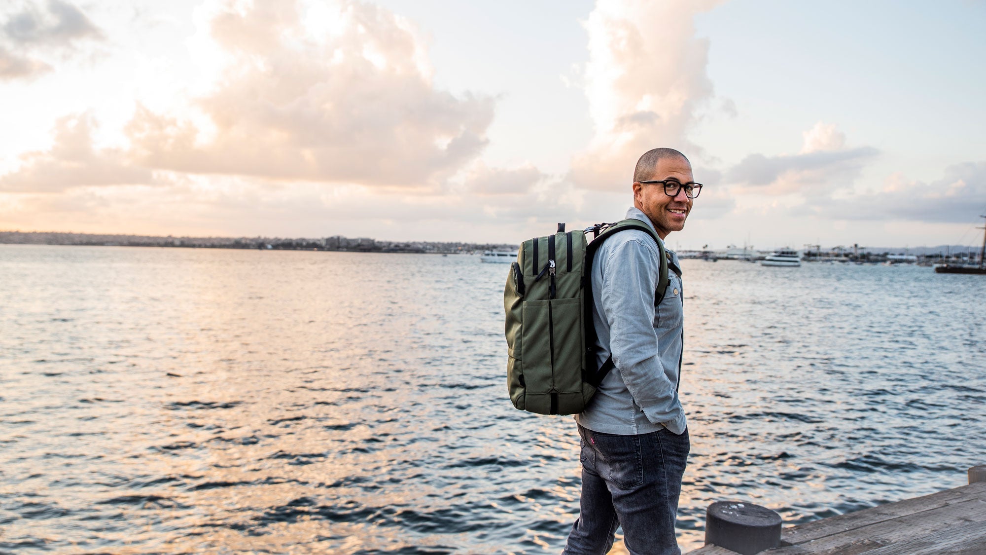 Man wearing glasses and a gray jacket with a green V1 Travel Backpack, smiling while standing on a wooden dock by the water at sunset with boats in the background.