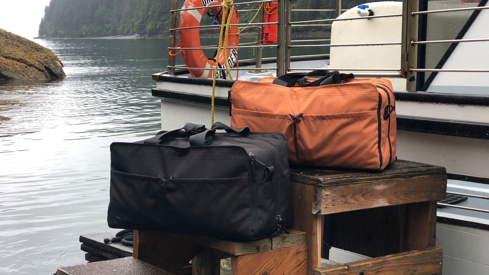 Two travel bags, one black and one orange, placed on wooden stools near a boat docked by a calm water body with forested shoreline in the background.