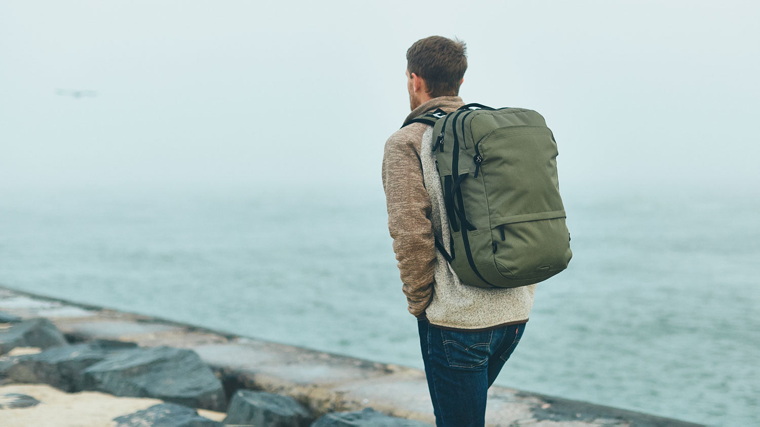 Man wearing a green V1 Travel Backpack standing on a rocky shore looking out over the water.