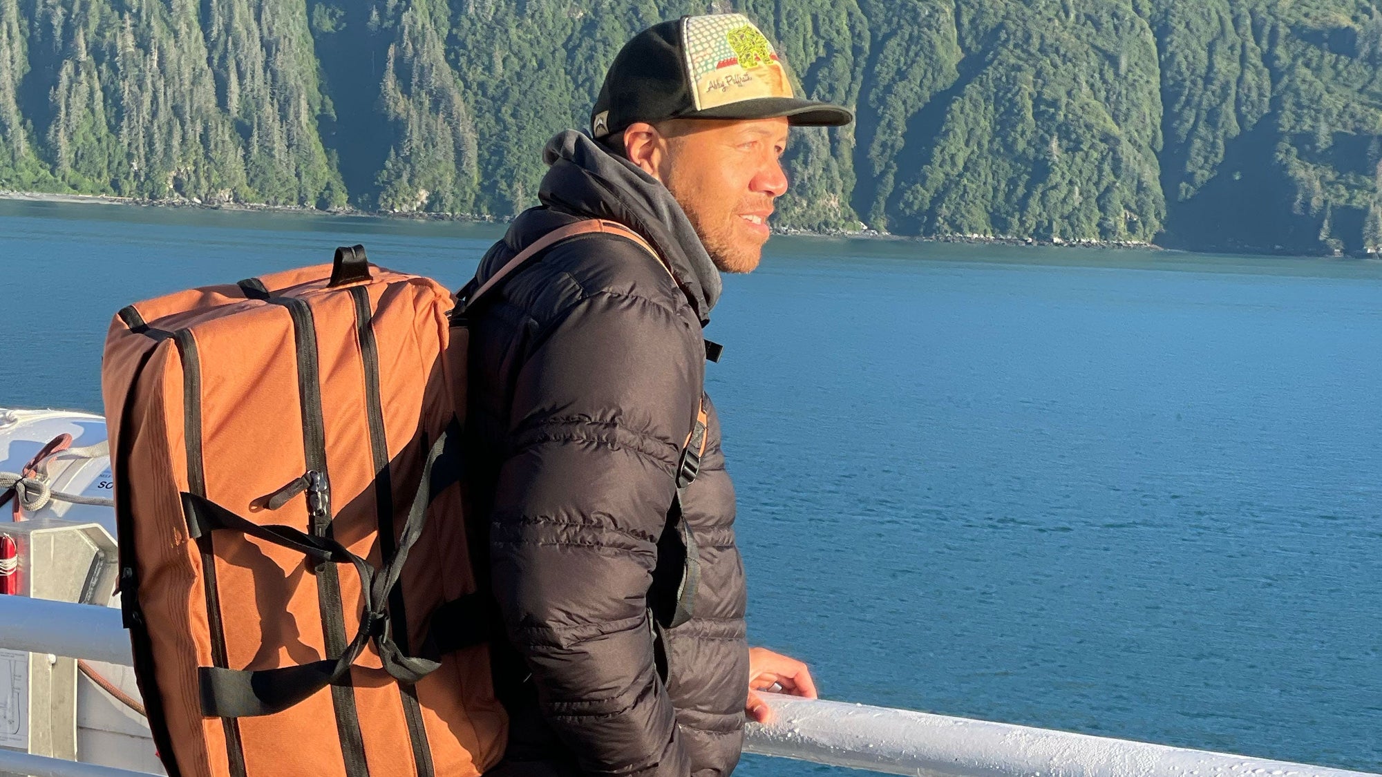 Man wearing a black puffer jacket and a cap with a large orange 50L Duffel, standing by a railing overlooking a blue body of water with forested cliffs in the background.