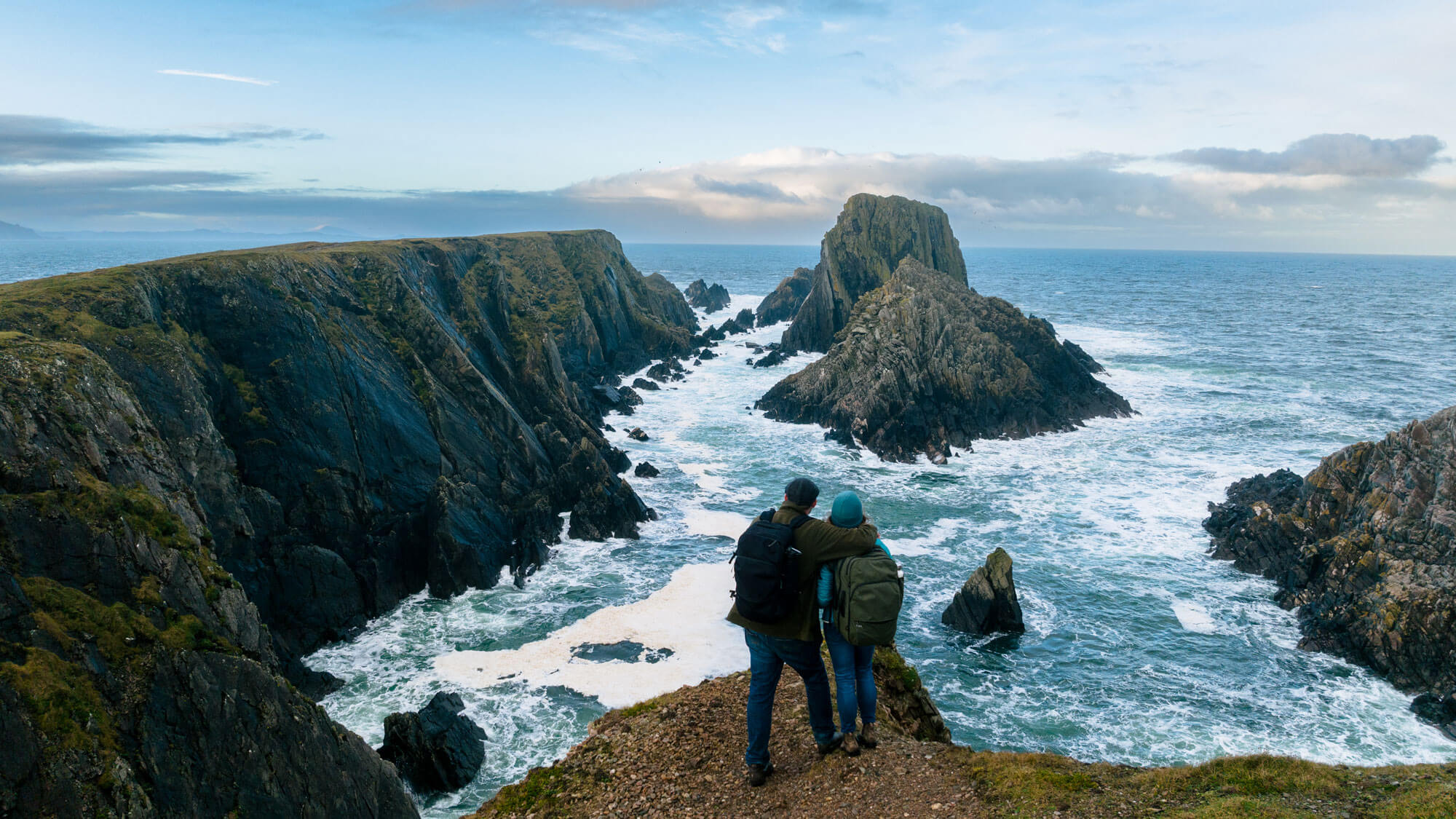 Two hikers with Travel Backpacks standing on a cliff overlooking rocky coastline and ocean waves in Ireland.
