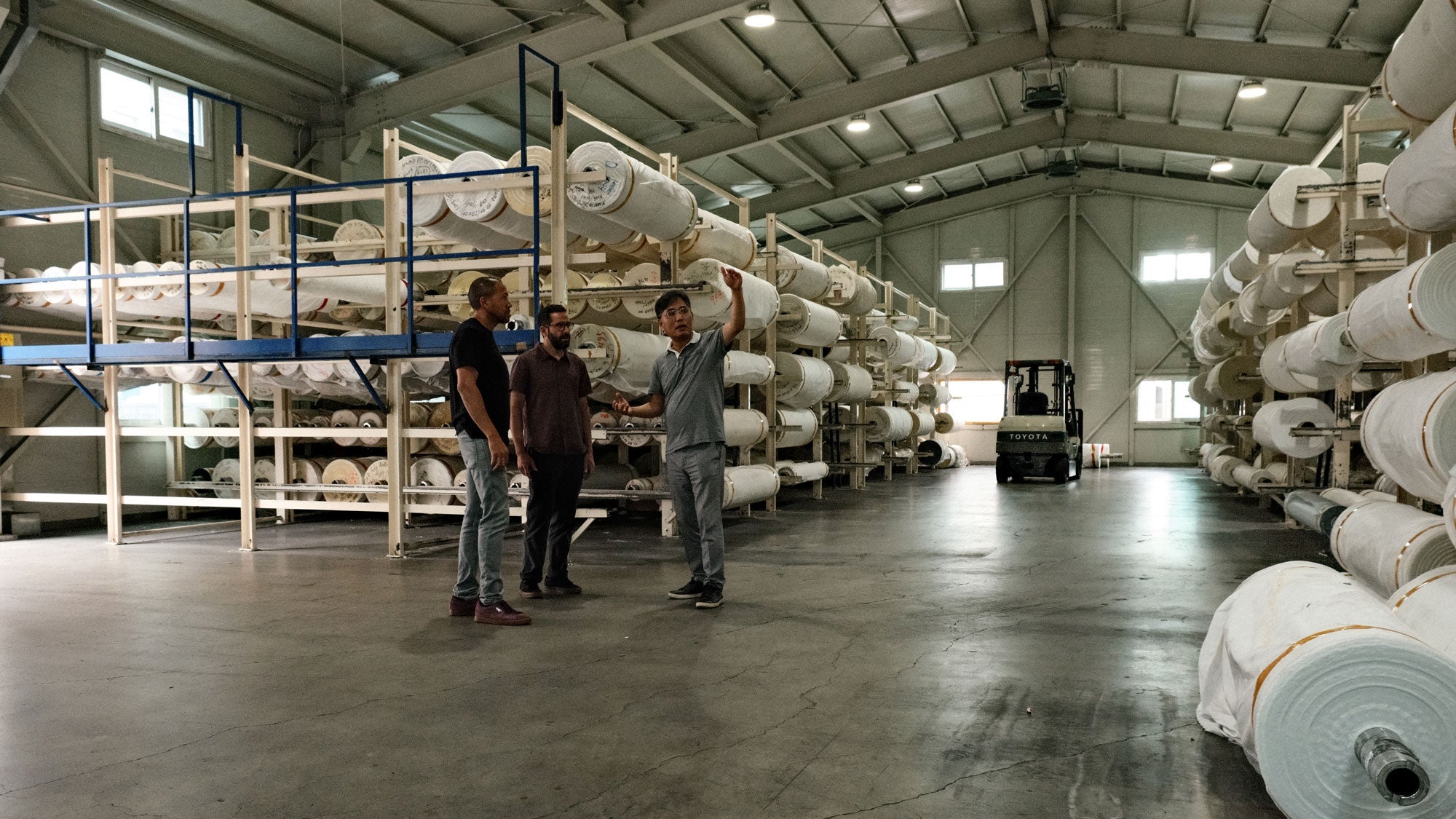 Three men standing in a fabric mill warehouse with large rolls of fabric stacked on racks and a forklift in the background.