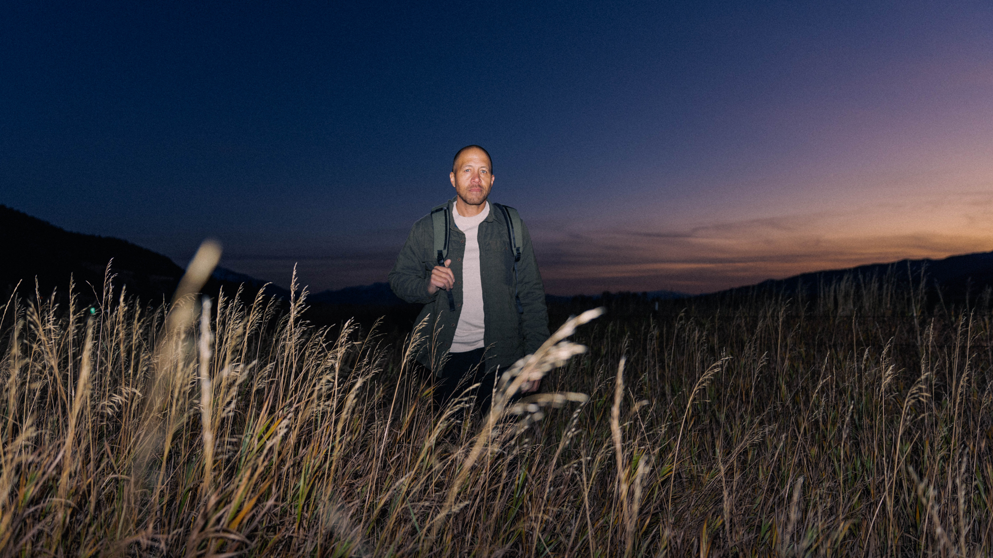 Pakt's founder holding a backpack standing in a field of tall grass at dusk with mountains in the background and a darkening sky.