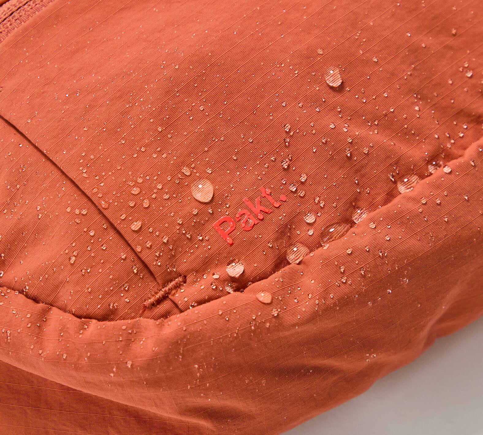 Close-up of an orange fabric with water droplets and the word "PAKT" embroidered on it, highlighting water resistance of the Stash Packable Sling 4L bag.