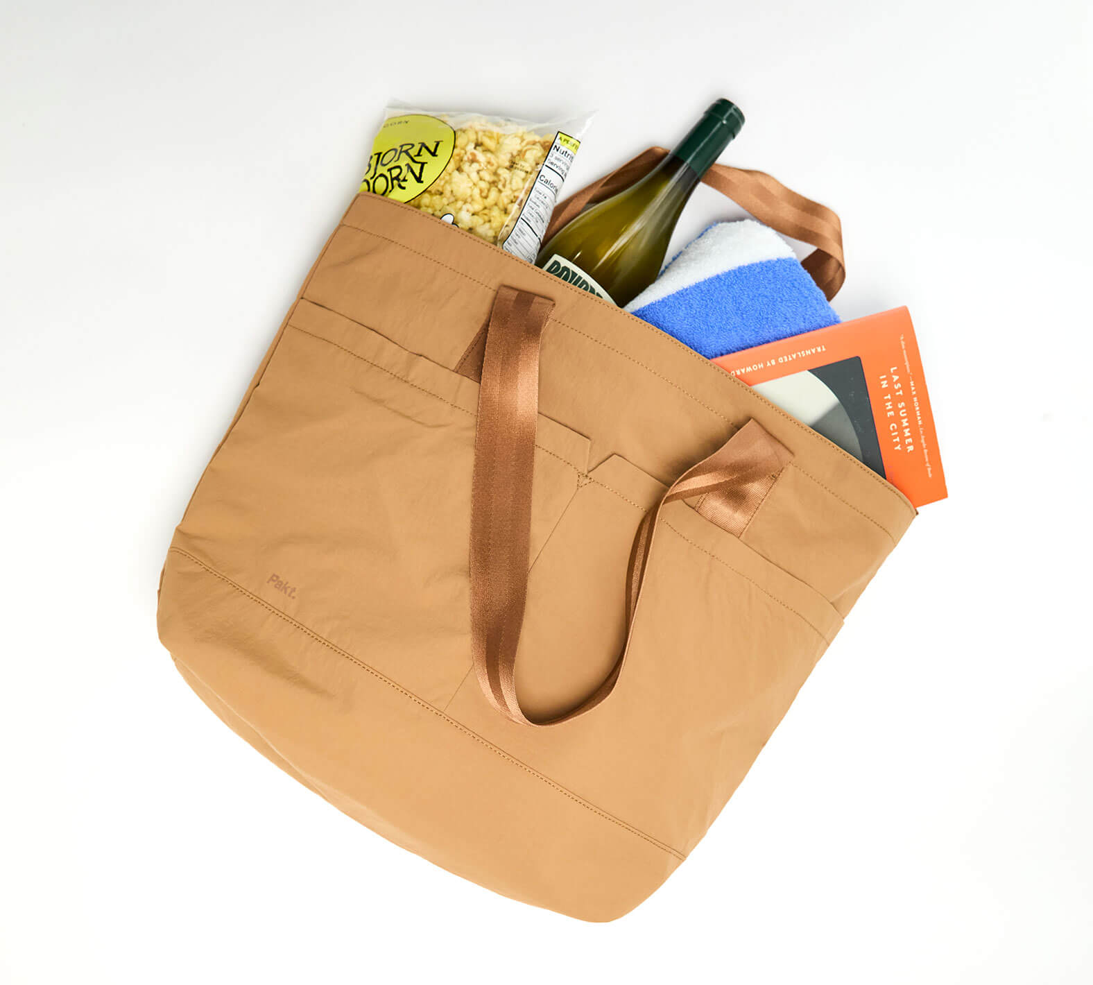 Brown tote bag with handles, containing a wine bottle, popcorn, a blue towel, and a book.