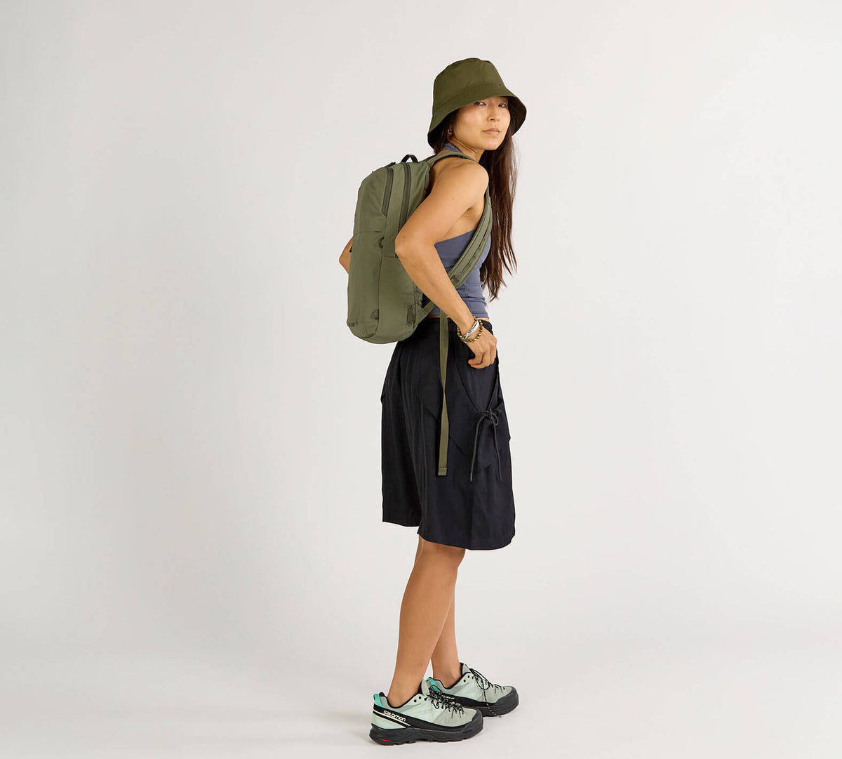 Woman wearing Stash Packable Backpack 16L in olive green, standing sideways in casual outfit.