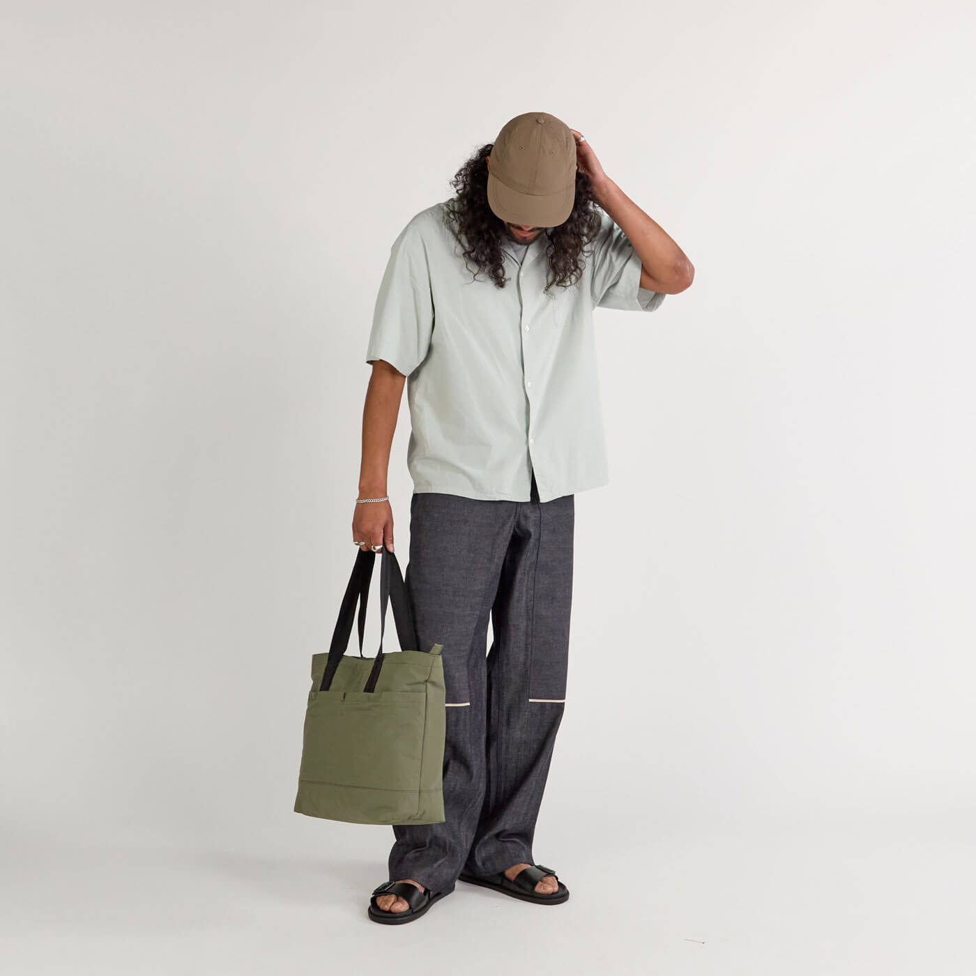 A person wearing a beige cap, light gray short-sleeve button-up shirt, dark gray pants with light stripes near the knees, black sandals, and holding a green Stash Tote bag with black handles, standing against a plain light background.