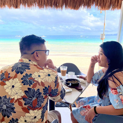 Couple seated at a beachfront table under a thatched roof, talking over iced drinks with ocean and sandy beach beyond.