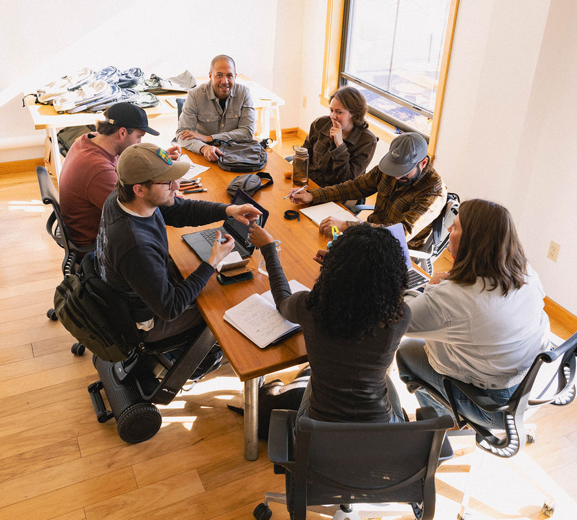 The Grandcart team of seven people collaborating around a wooden table in a bright room, some using laptops and notebooks.