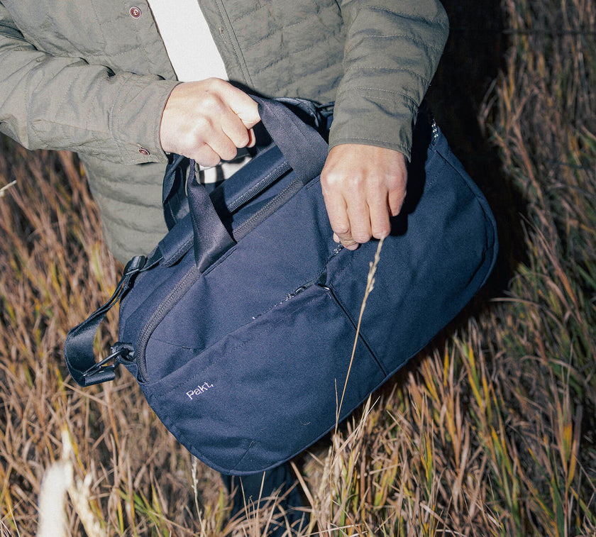 Person holding a black 25L Travel Duffel, outdoors in tall grass