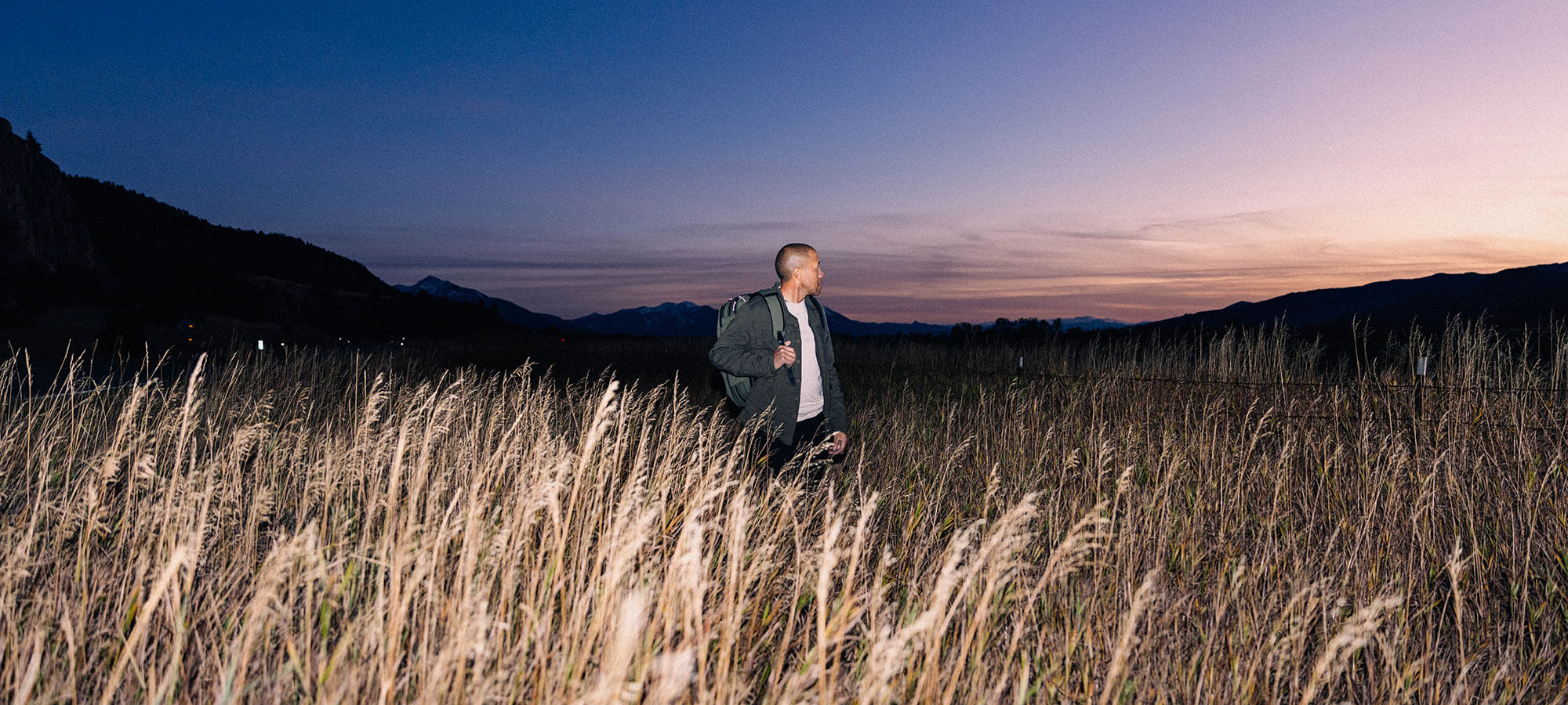 Pakt's Founder wearing a Travel Backpack standing in a tall grassy field at dusk with mountains in the background under a purple and blue sky.