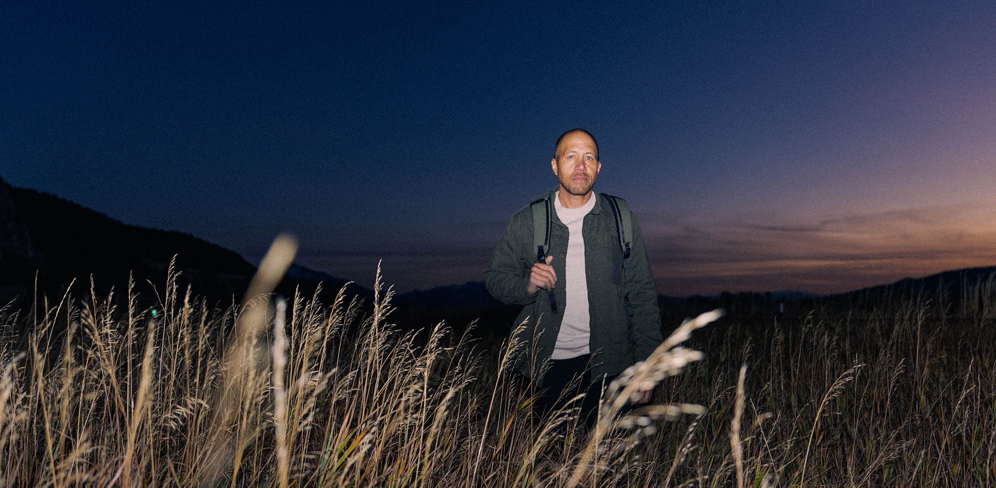 Man standing in a field of tall grass at dusk wearing a backpack and jacket