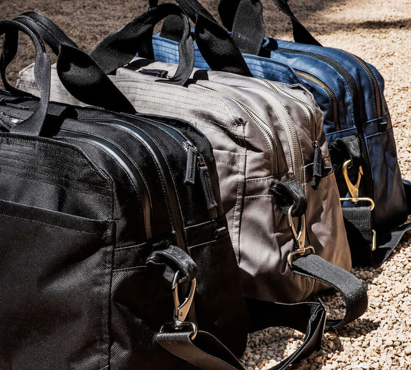 Three V1 Grandcart Ones in black, gray, and blue with zippers and shoulder straps placed on gravel ground.