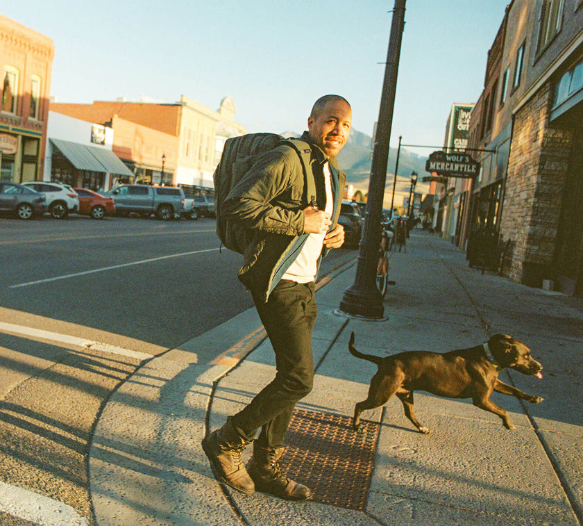 Pakt's founder wearing a Travel Backpack walking a black dog on a city sidewalk near storefronts including a sign for Wolf's Mercantile.