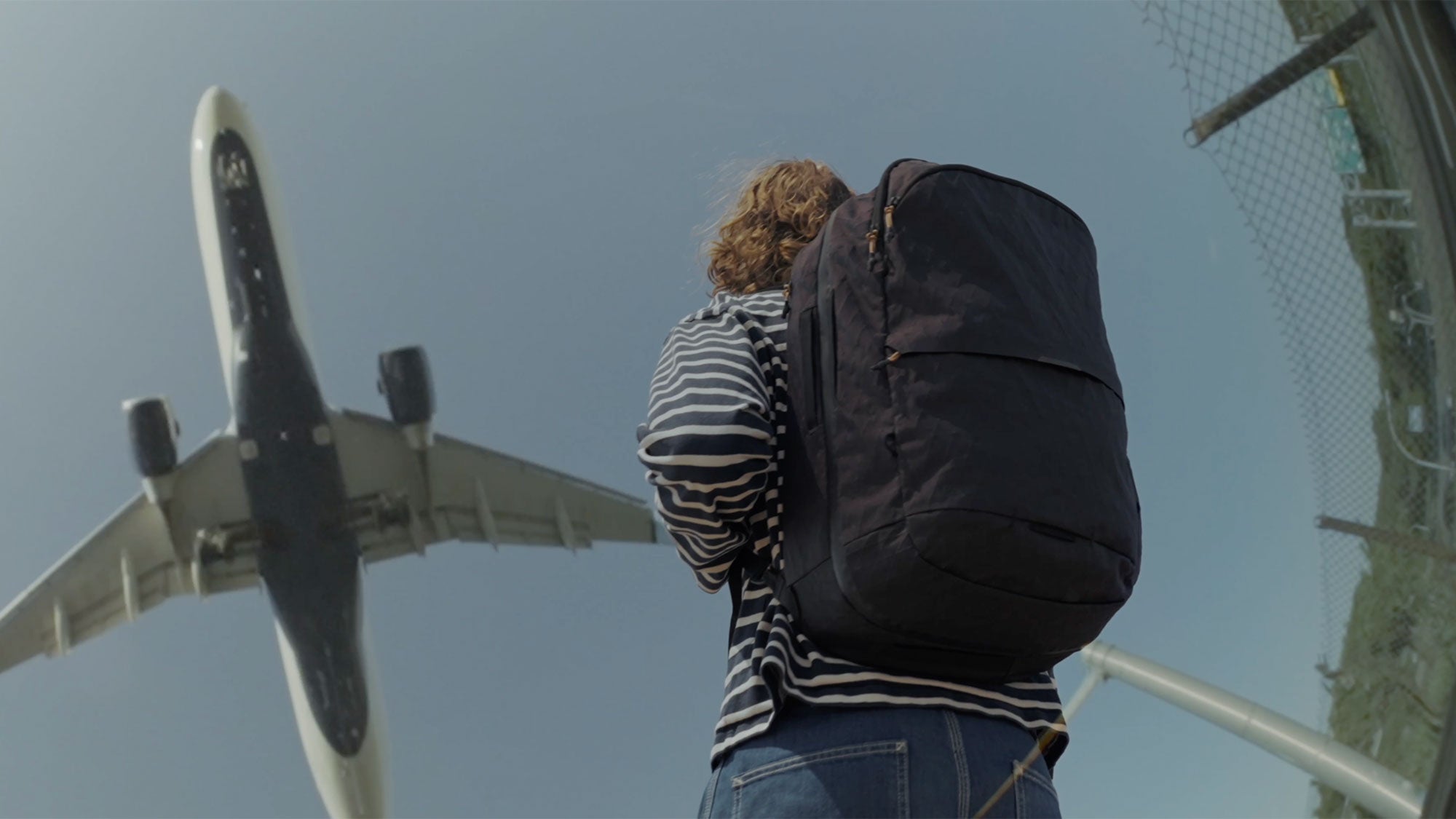 Person wearing a large black V1 Travel Backpack looking up at a plane flying overhead against a clear sky near a chain-link fence.