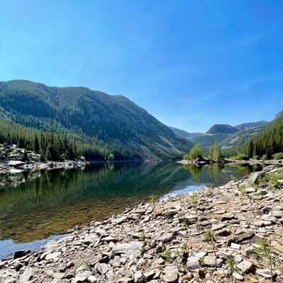 Rocky lakeshore with clear alpine lake reflecting forested mountains under a bright blue sky