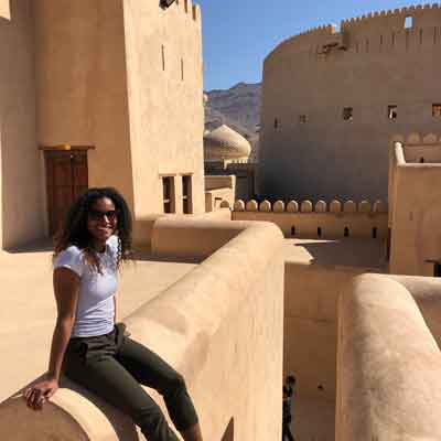 Woman wearing sunglasses and white top sitting on a mud‑brick fort wall, beige desert fortress and domed tower in background.