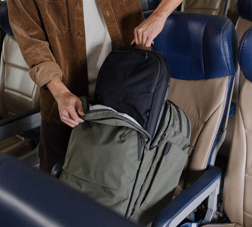 Person placing a smaller Everyday 15L Bag inside a larger green Travel Backpack on an airplane seat