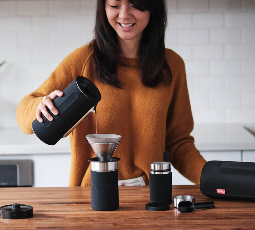 Woman pouring water from a black kettle into a coffee dripper on a wooden table with coffee-making accessories nearby.