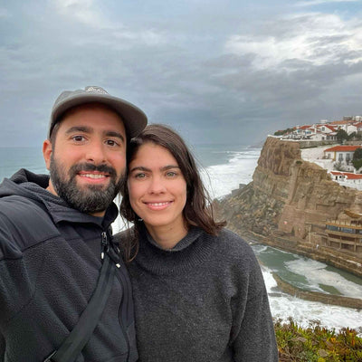 Smiling couple selfie on a coastal cliff with waves below and white houses on the cliff under a cloudy sky