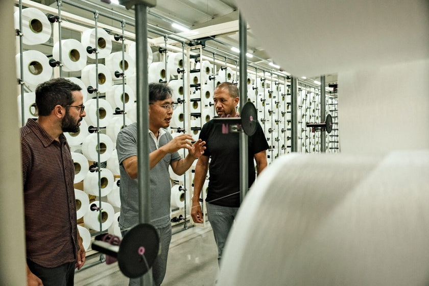 Three men discussing in a factory with large spools of white material on racks behind them.