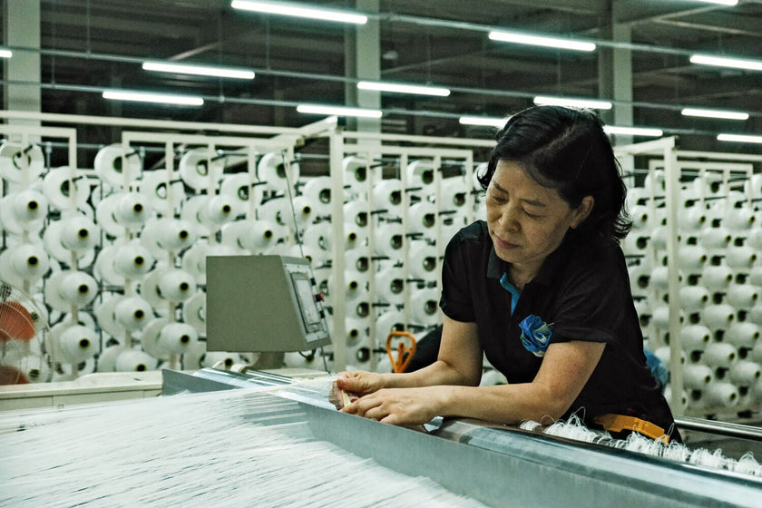 Woman inspecting threads on a textile machine in a factory with rows of yarn spools in the background