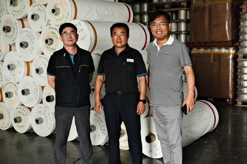 Three men standing in a warehouse in front of large rolls of white material, two wearing black shirts and one in a gray polo shirt, smiling and posing for the photo.