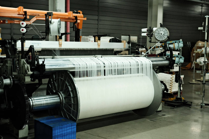 Industrial textile weaving machine producing white fabric on a large roll in a factory setting