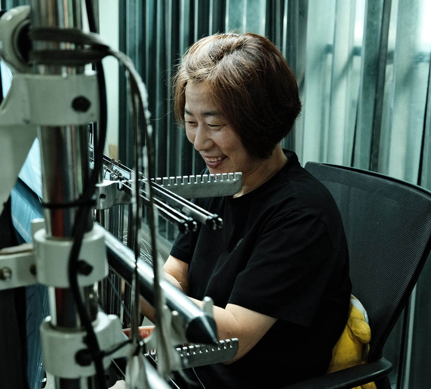 Woman operating a textile weaving machine, smiling while seated in a chair.