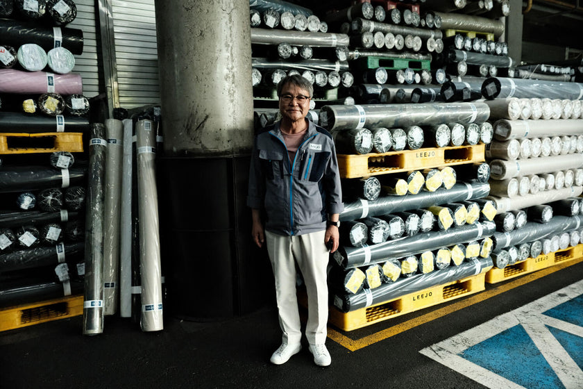 An older person wearing glasses and a gray jacket stands in a warehouse surrounded by stacks of rolled fabric on yellow pallets labeled 'LEEJO'.