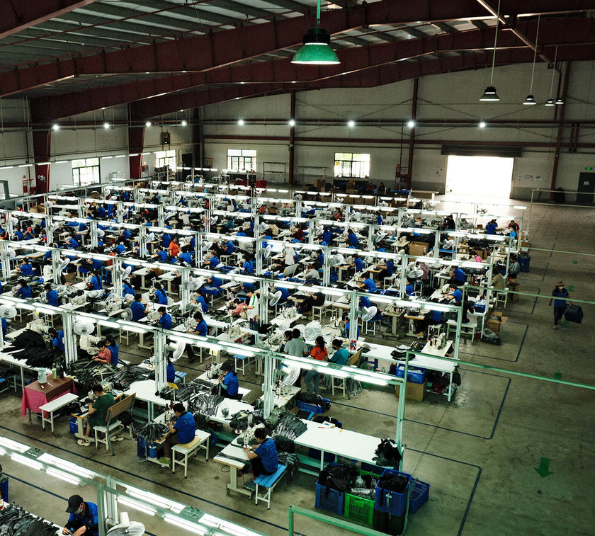 Large factory floor with rows of workers seated at sewing machines assembling products under bright overhead lights.