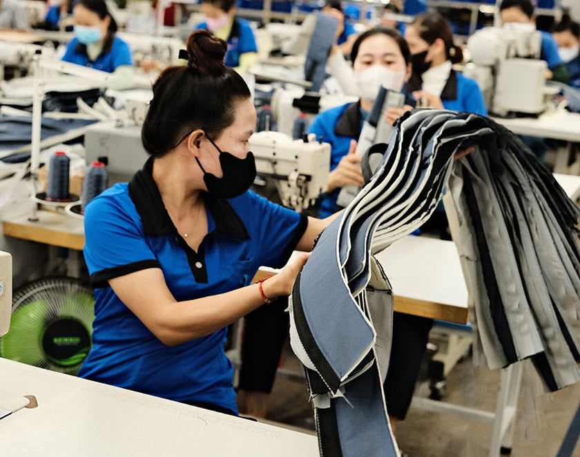 Woman in a blue shirt and black mask handling fabric pieces in a garment factory with other masked workers sewing in the background.