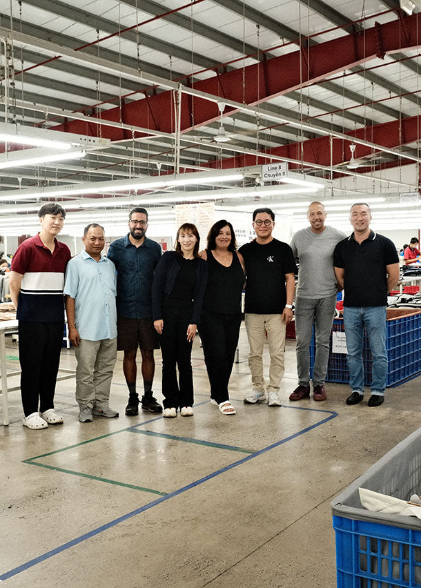 Group of eight people standing in a warehouse or factory with industrial lighting and storage bins in the background.