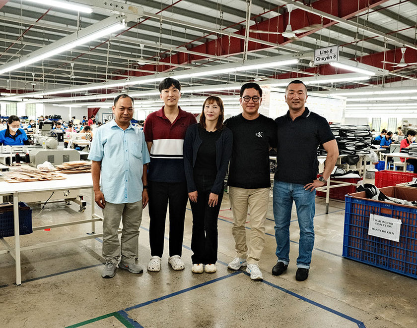 Five factory workers standing together in a manufacturing workshop with sewing machines and other workers in the background.